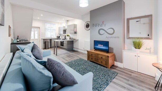 A modern living room with a sofa, a TV, and a kitchen in the background. - Infinity Apartments Harrow Road (Liverpool)