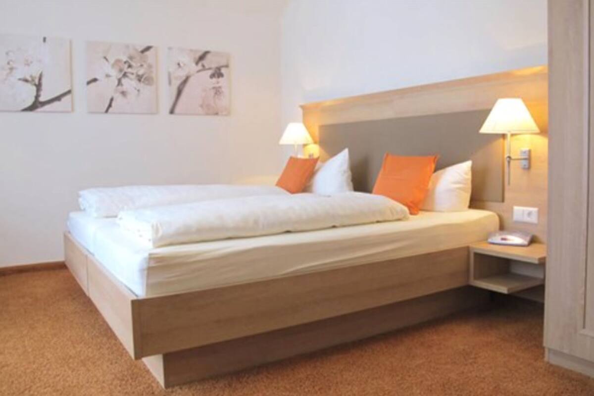 Een tweepersoonsbed met wit en oranje beddengoed, twee bedlampen en drie kunstwerken aan de muur.