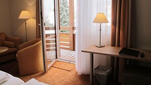 Une chambre d’hôtel avec un lit, un bureau doté d’une lampe, une chaise et une vue sur une terrasse en bois et des arbres à l’extérieur.