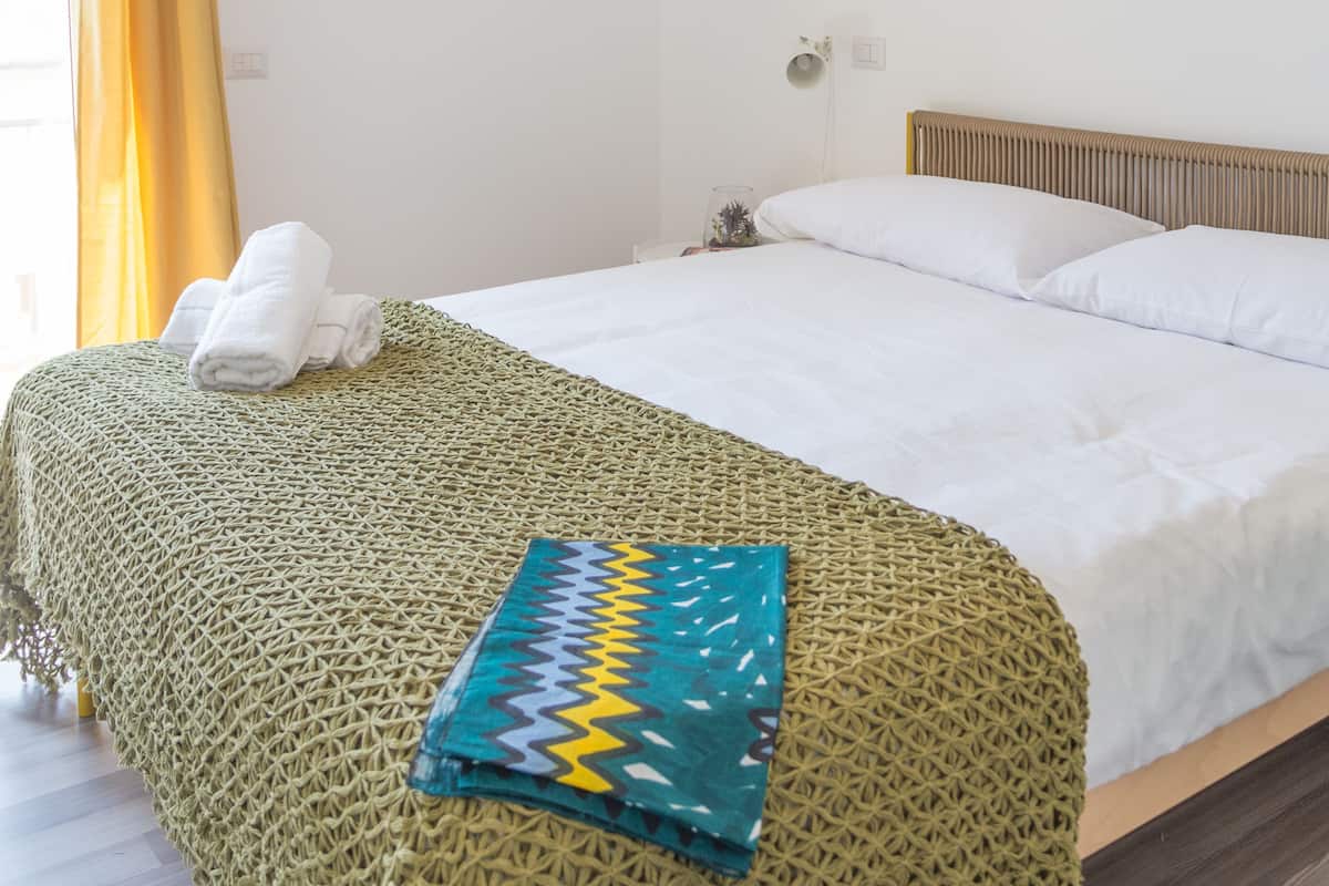 Een net opgemaakt bed met een groene deken, een hoofdbord en een aan de muur bevestigde airconditioner.