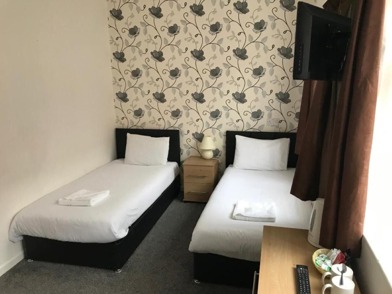 Habitación de hotel con dos camas individuales, una mesita de noche, televisor y ventana con cortinas.