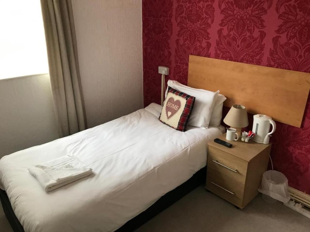A neatly made bed with a heart-shaped pillow, a bedside table with a lamp, a kettle, and a remote control.