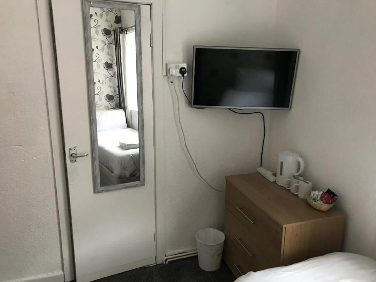 A hotel room with a bed, a wooden dresser, a television mounted on the wall, a window with curtains, and a trash bin.