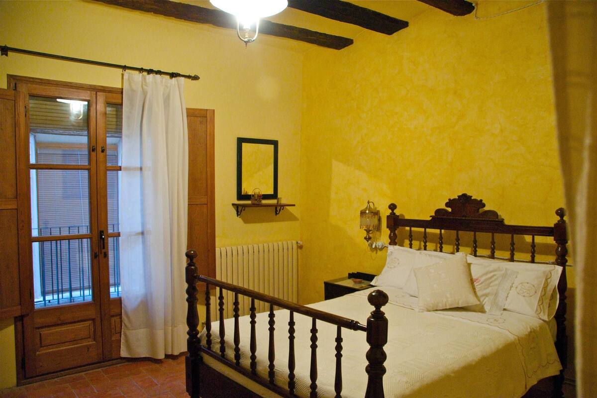A bedroom with a wooden bed, a nightstand, a wall-mounted lamp, and a window with a curtain.