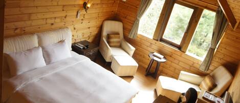 Habitación de cabaña de madera con cama, dos sillones, una mesita y ventana con vista a follaje.
