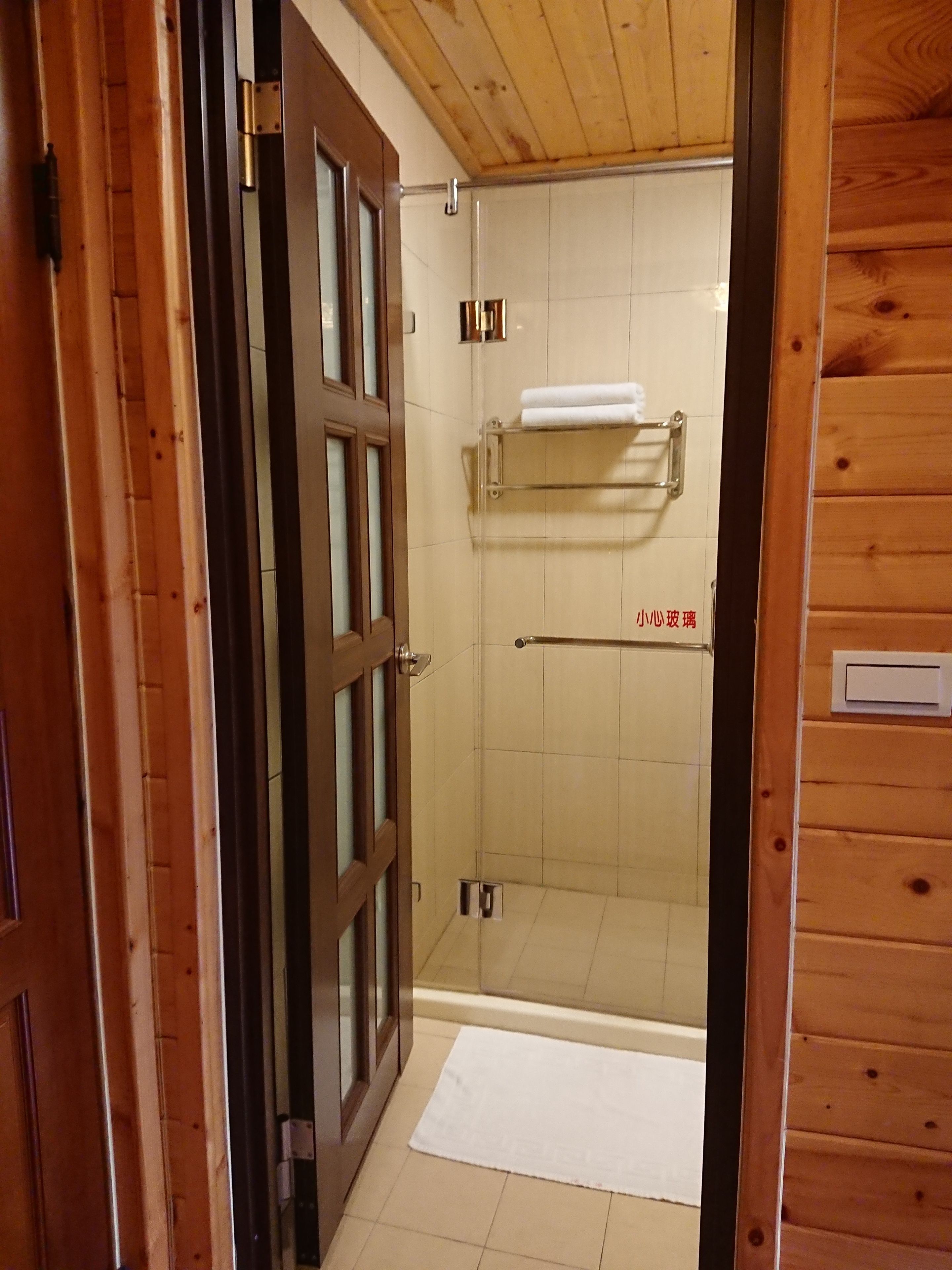 A wooden-paneled bathroom with a glass shower enclosure, a towel rack, and a white mat on the floor.