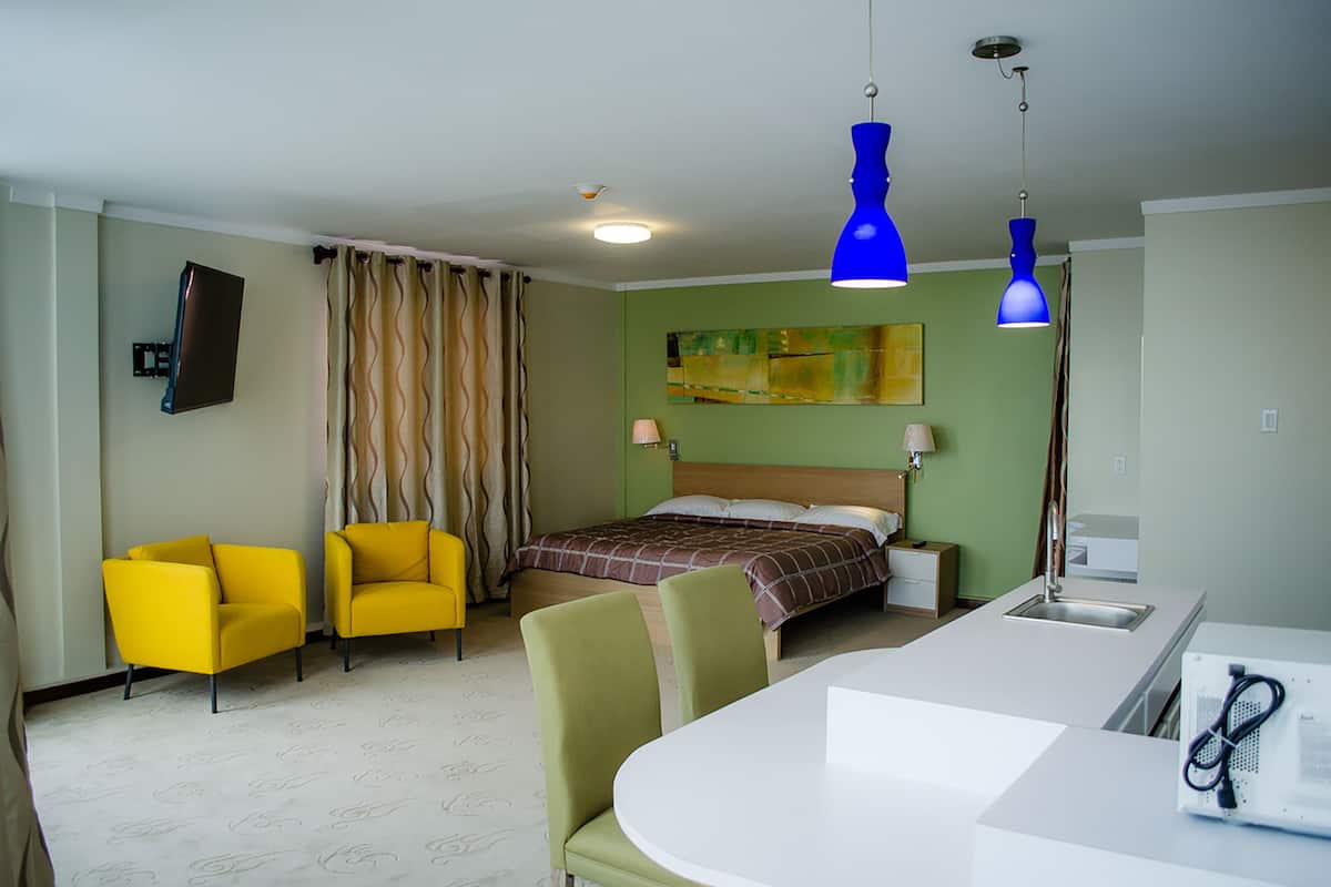 Una habitación de hotel con una cama, dos sillas amarillas, una mesa blanca y una lámpara colgante azul.