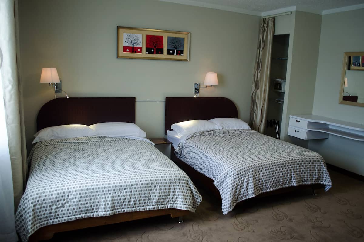 Habitación de hotel con dos camas, cada una con una colcha blanca y gris estampada, un cabecero de madera, una mesita de noche, una lámpara y un cuadro enmarcado en la pared.