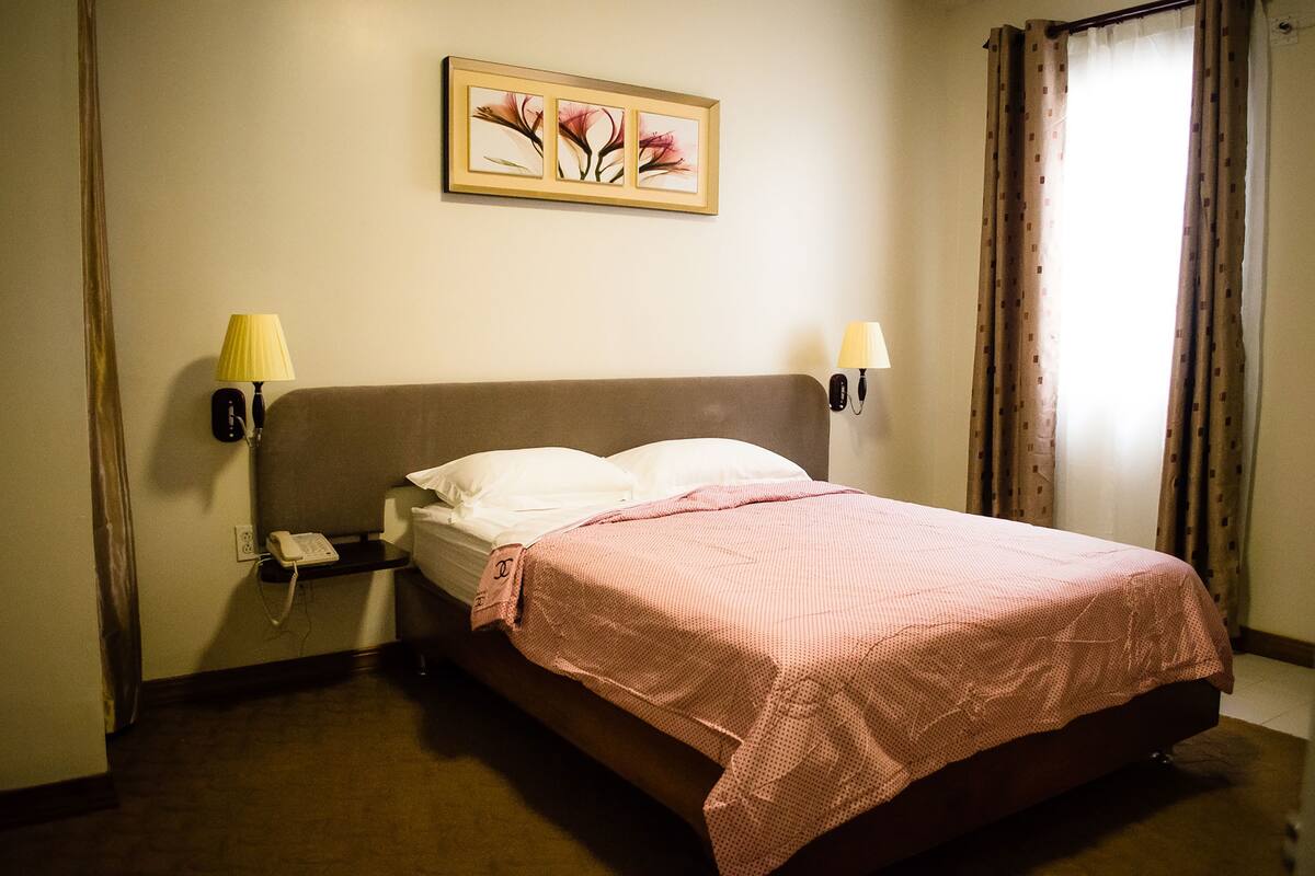 Habitación de hotel con cama, mesita de noche con lámpara, teléfono y un cuadro en la pared.