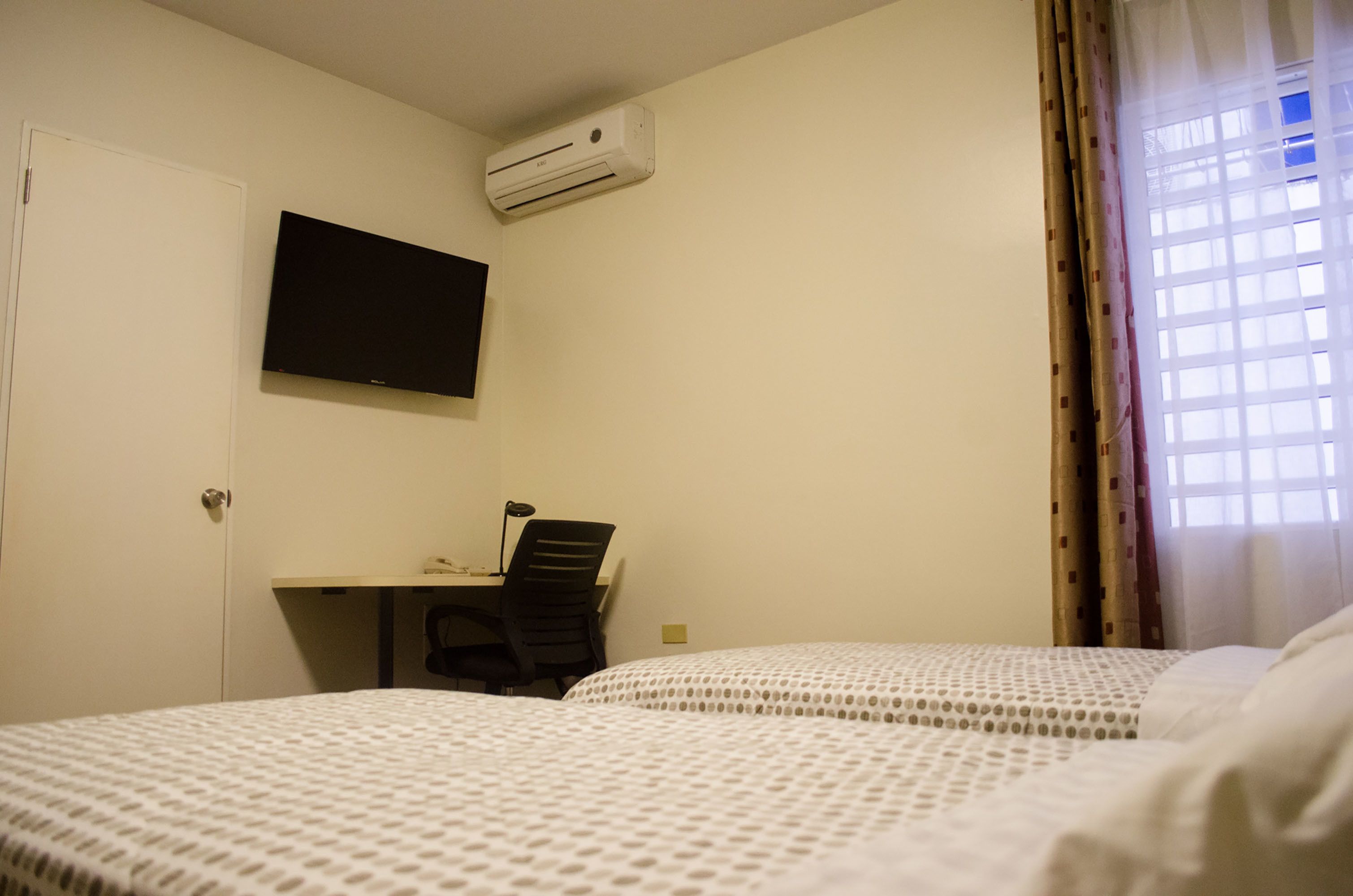 A hotel room with two beds, a desk with a chair, a TV mounted on the wall, and an air conditioning unit.