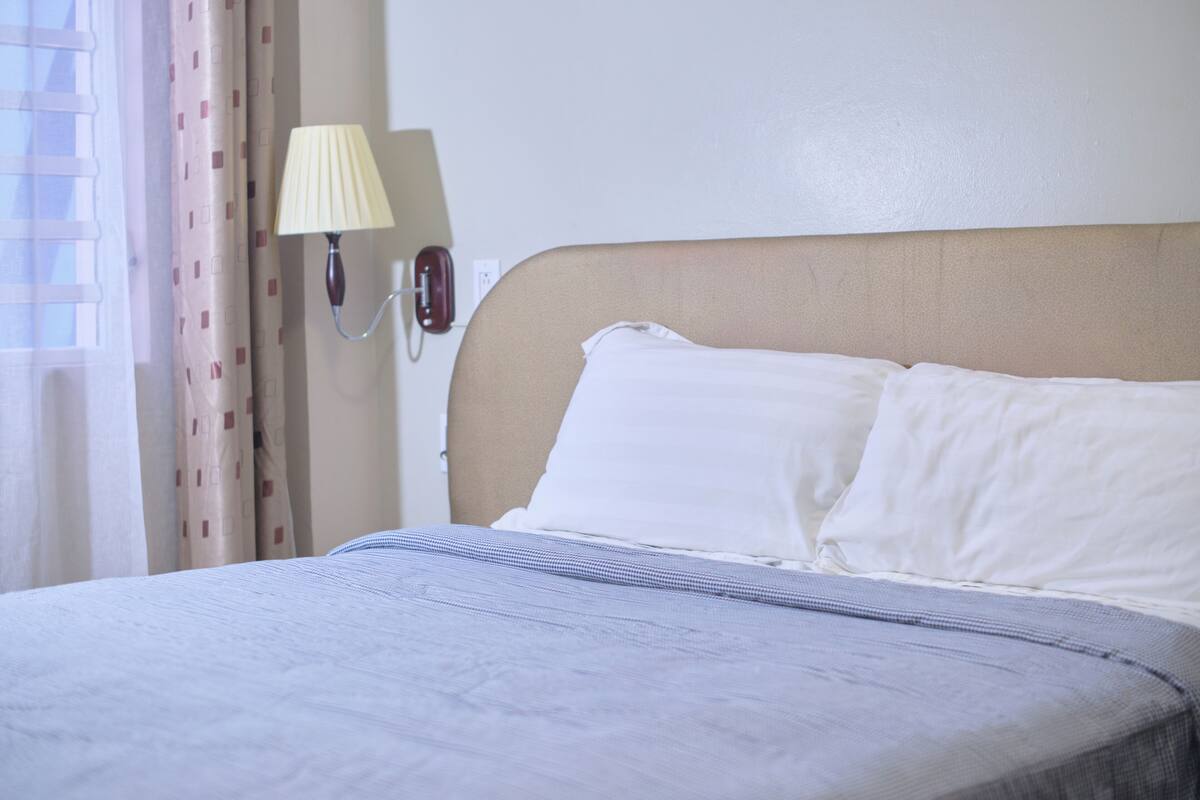Una cama bien tendida con sábanas blancas, cabecera color beige y una lámpara fijada a la pared.