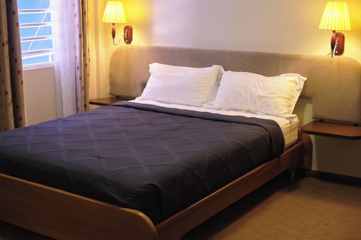 Una cama bien hecha con un edredón oscuro, dos lámparas de noche y una ventana con cortinas.