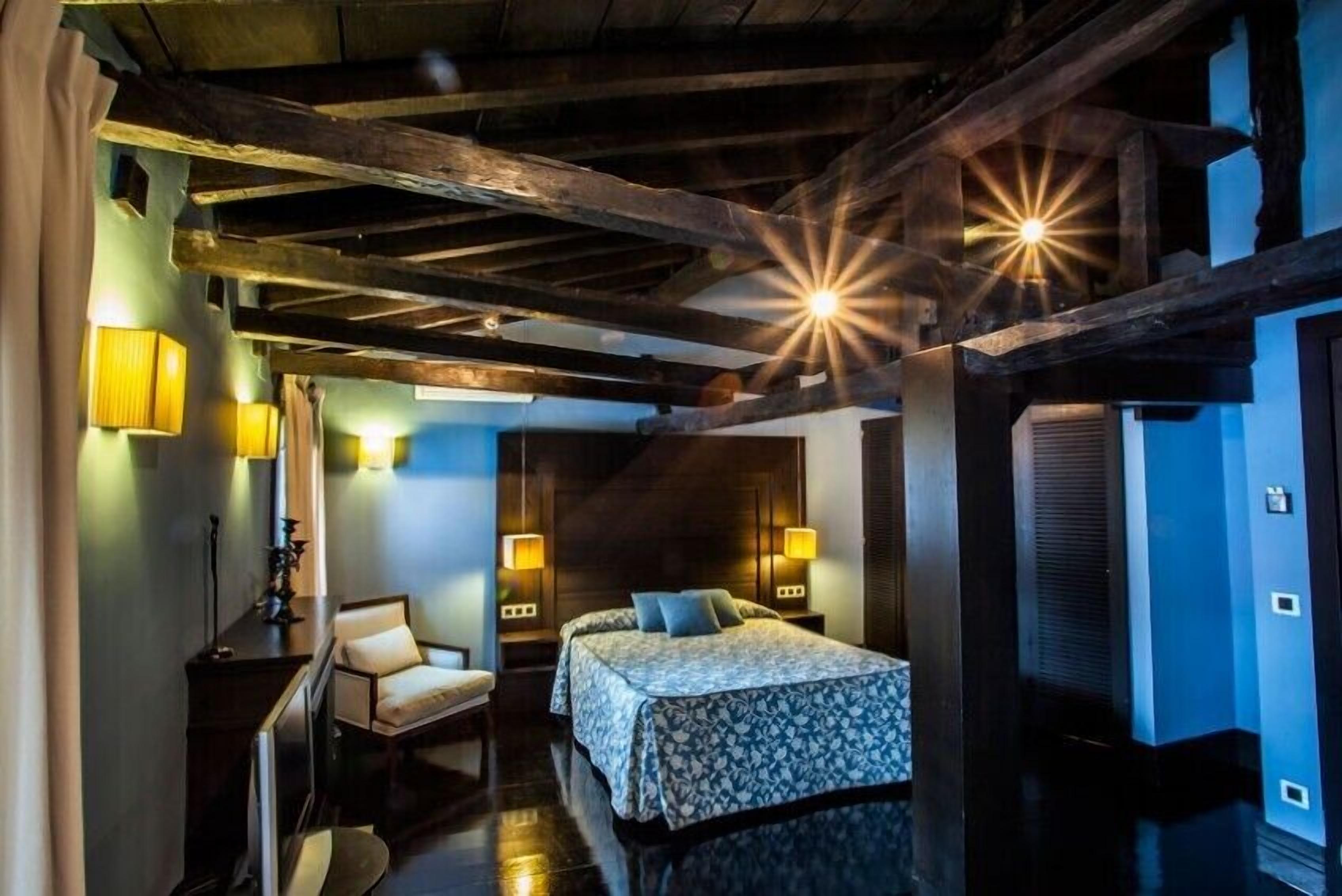 Une pièce faiblement éclairée, avec des poutres apparentes, un lit recouvert d’une couvre-lit à motifs et des lampes fixées au mur.