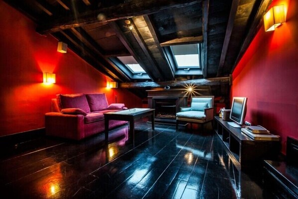 Un acogedor dormitorio en ático con pared roja, un sofá, una silla, una mesita y un televisor.