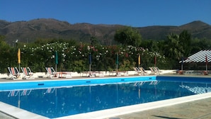 Una piscina con sillones de descanso y sombrillas, rodeada de vegetación y montañas al fondo.