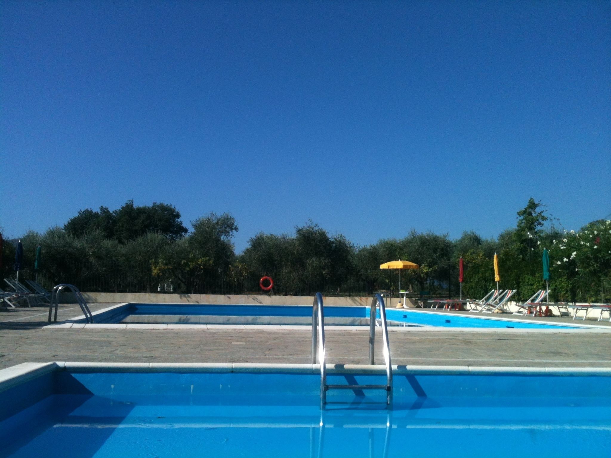 Una piscina con escalera, sillas de descanso y sombrillas.