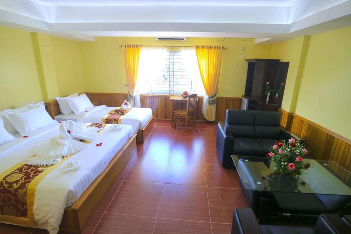 Une chambre d’hôtel avec deux lits, un canapé, une petite table ornée d’un vase de fleurs et une fenêtre avec des rideaux.