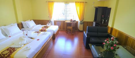 Quarto de hotel com duas camas, um sofá, uma mesa pequena com um vaso de flores e uma janela com cortinas.