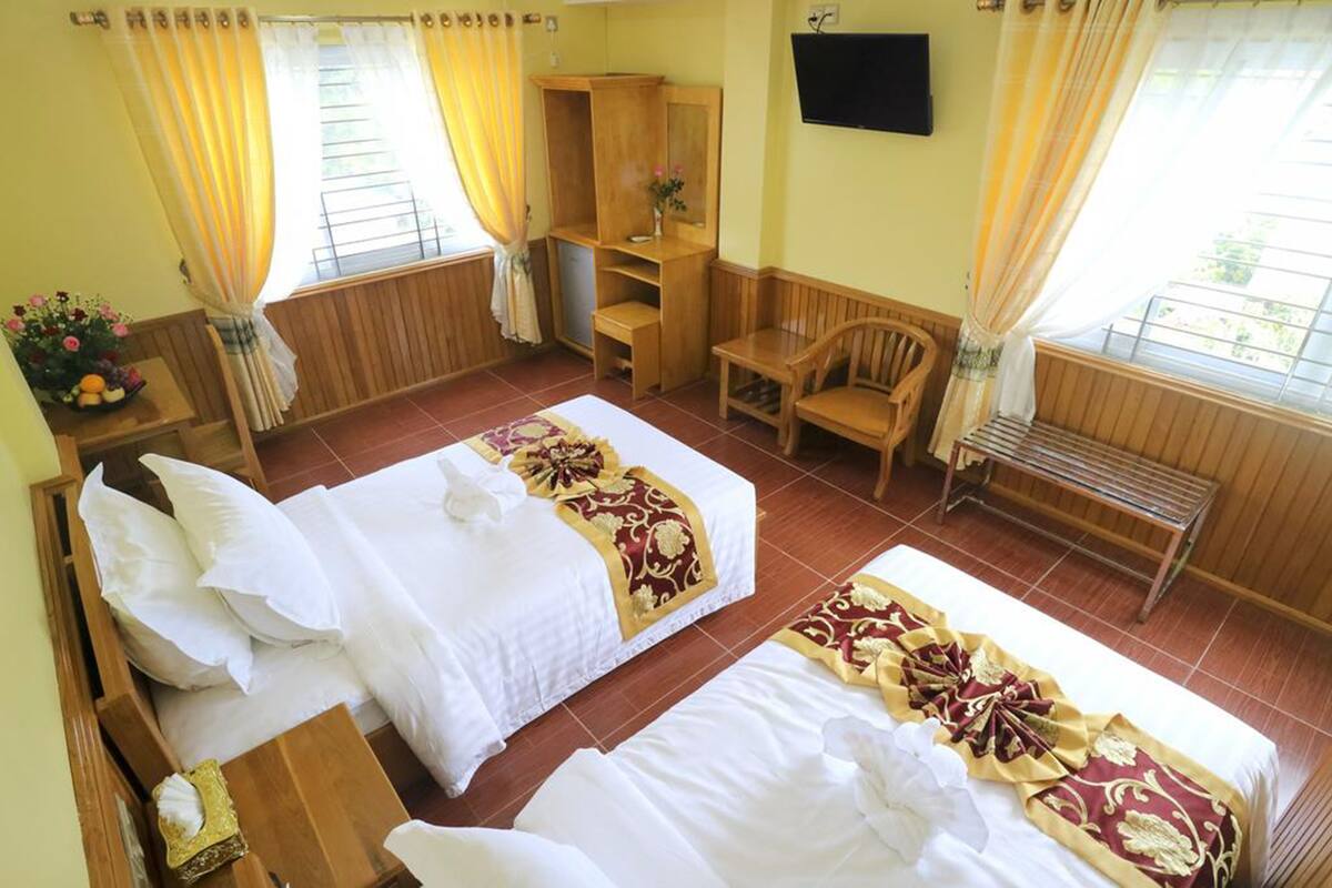 Une chambre d’hôtel avec deux lits, un banc en bois, une télévision et une fenêtre avec des rideaux.