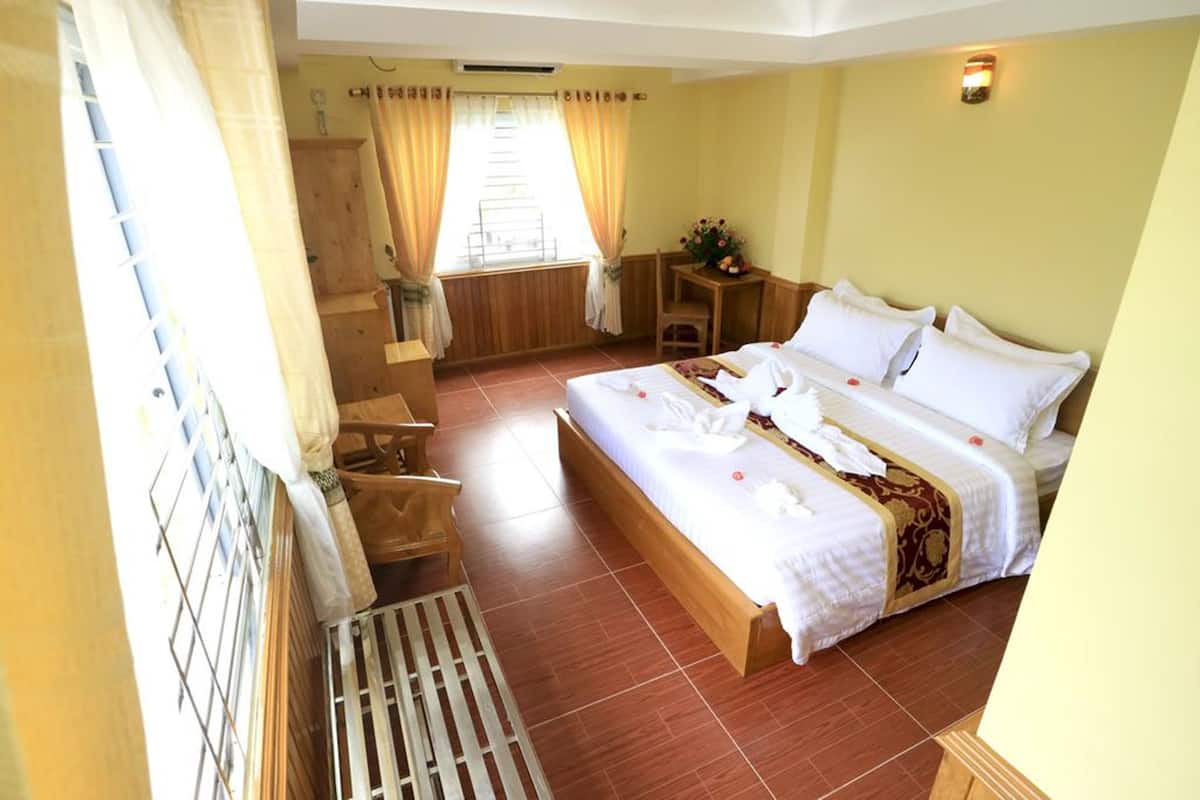 Une chambre d’hôtel avec un grand lit, des meubles en bois, une petite table avec un vase et une fenêtre avec des rideaux.