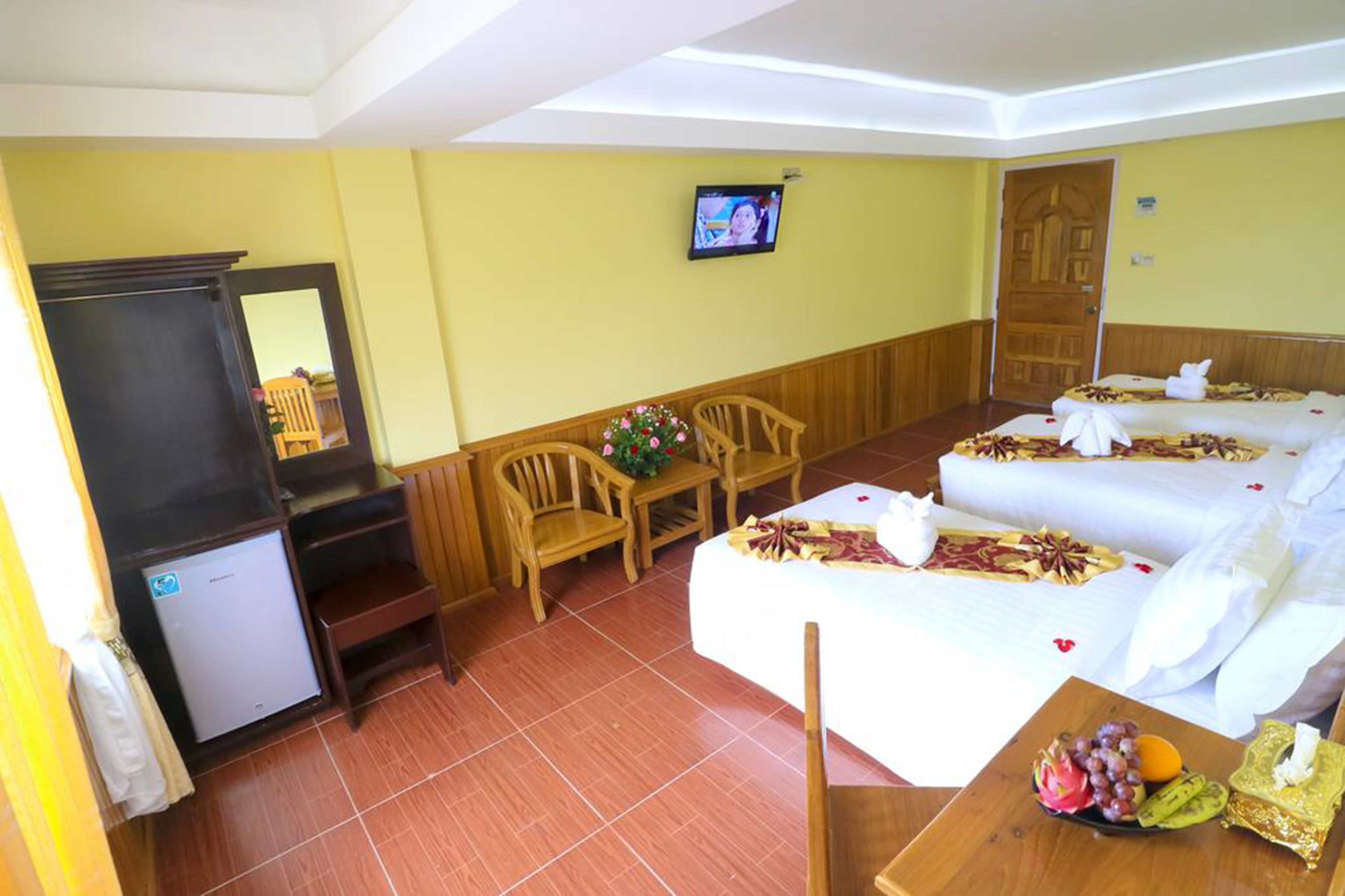 A hotel room with a bed, a TV, a small fridge, a wooden chair, a table with fruit, and a mirror.