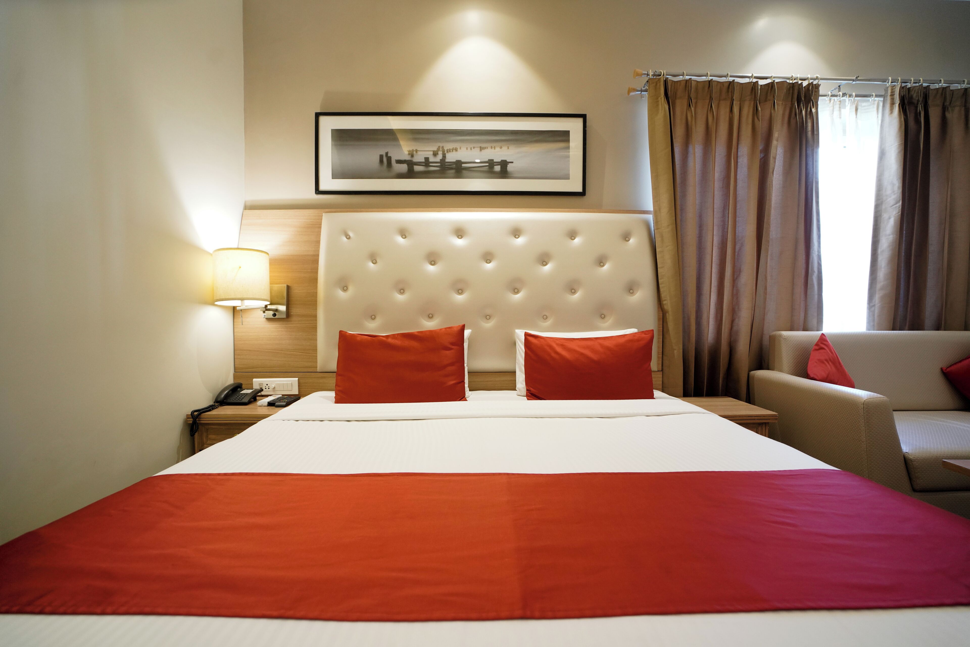 A hotel room with a large bed, a headboard, a bedside table with a lamp, a chair, and a framed picture on the wall.