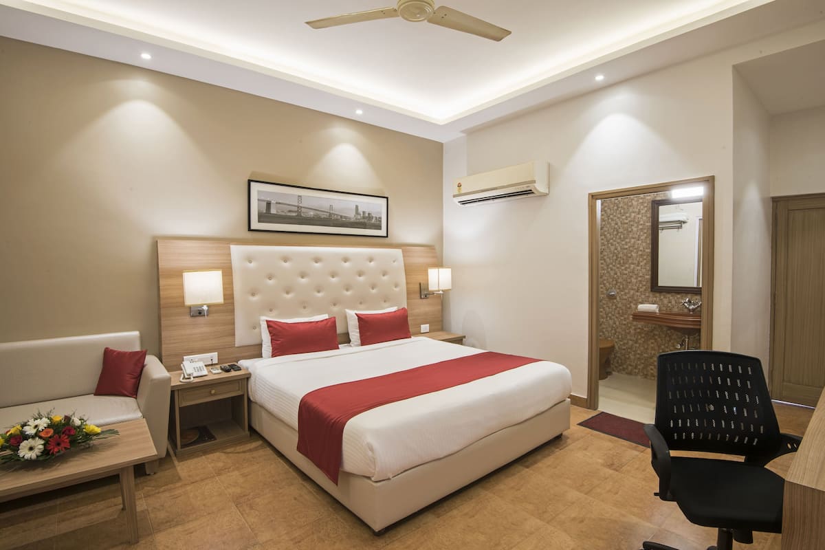 A hotel room with a large bed, a desk, a chair, a nightstand, a lamp, a picture on the wall, an air conditioning unit, and a bathroom door.