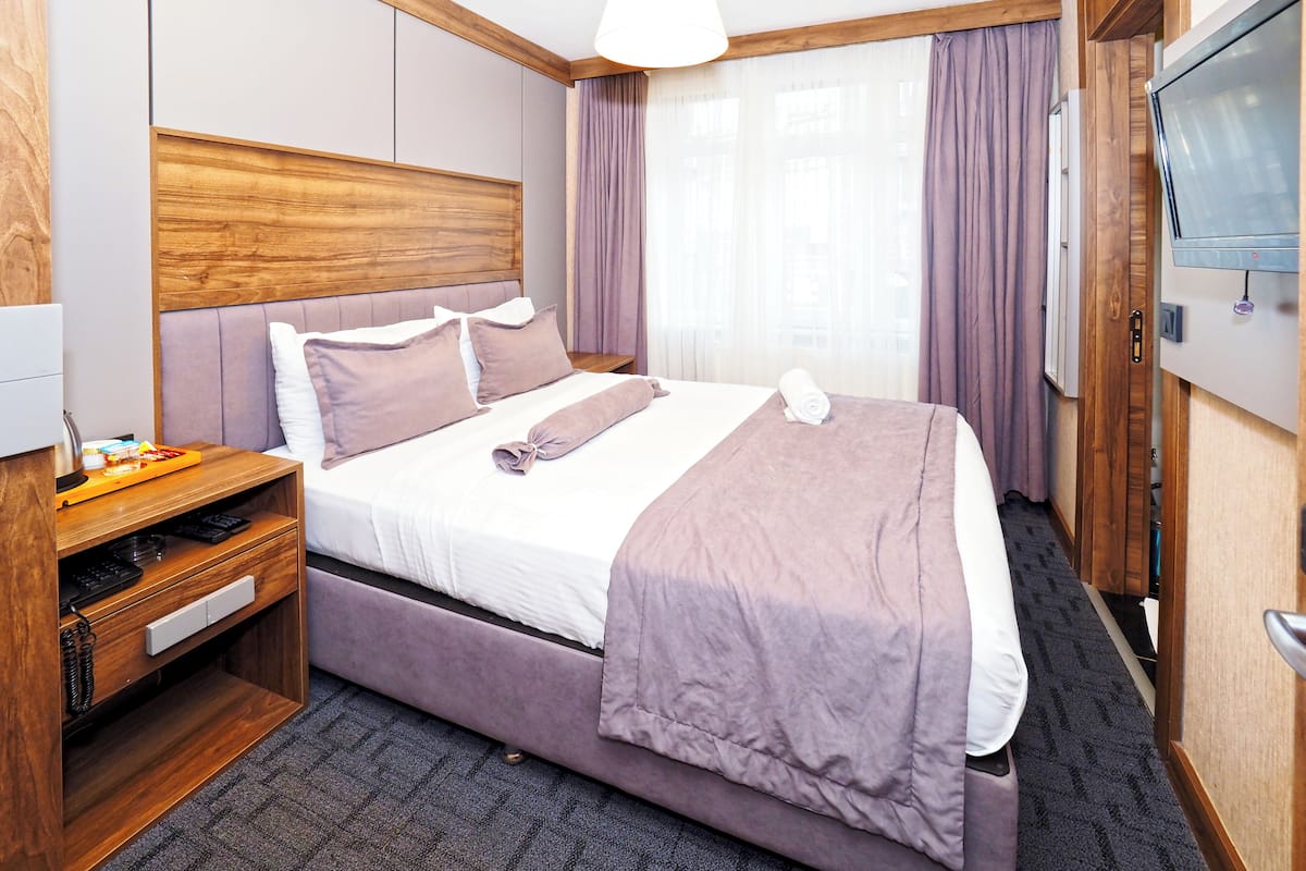 A hotel room with a large bed, wooden headboard, a bedside table with a phone and charger, a TV mounted on the wall, and a window with curtains.