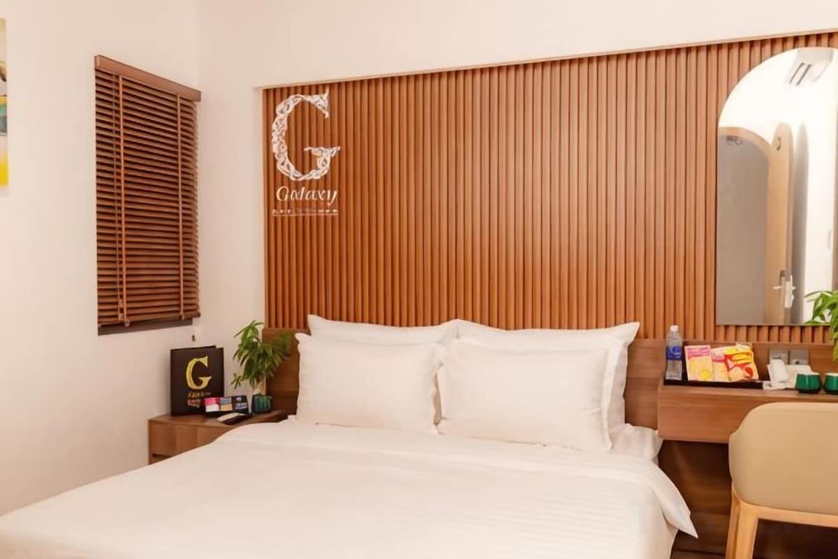 A hotel room with a bed, wooden headboard, a bedside table with a plant, a mirror, and a desk with toiletries.