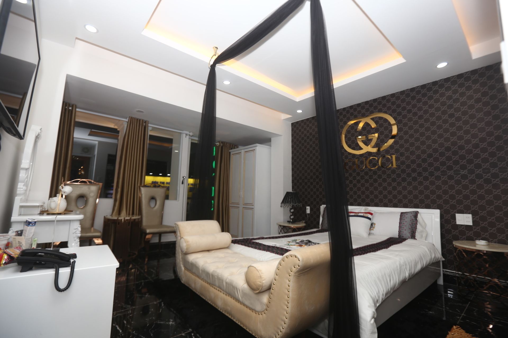 Een moderne hotelkamer met een hemelbed, een zithoek met een bank, een kaptafel met spiegel en een muur met een Gucci logo.