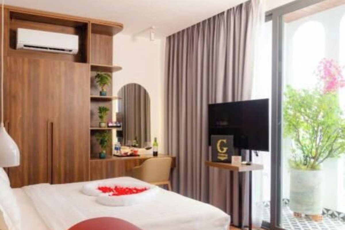 Una camera d'albergo con un letto, una TV, una scrivania con un computer, una finestra con tende e un balcone con una pianta.