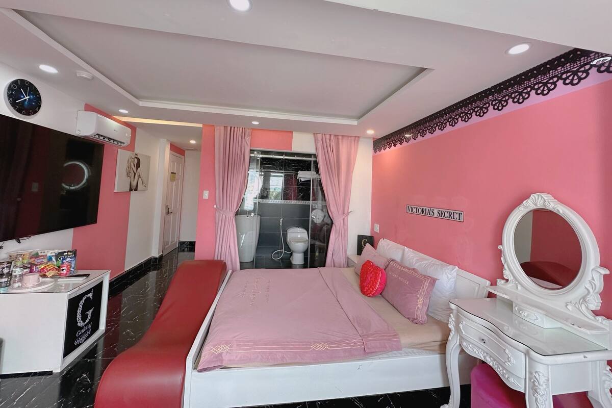 Een slaapkamer met een bed, een kaptafel met spiegel, een rode bank, een wandklok, en een bordje met de tekst 'VICTORIA'S SECRET'.