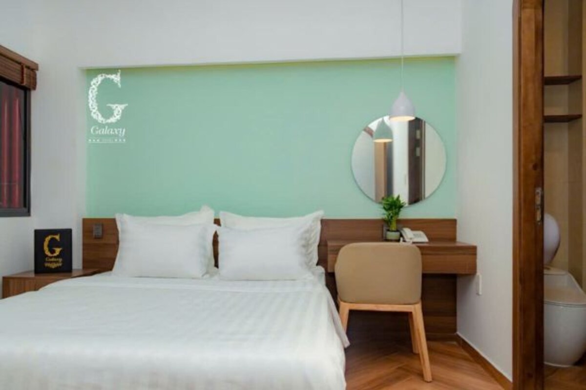 Una camera d'albergo con un letto, una scrivania, una sedia, uno specchio e un cartello incorniciato appeso al muro.