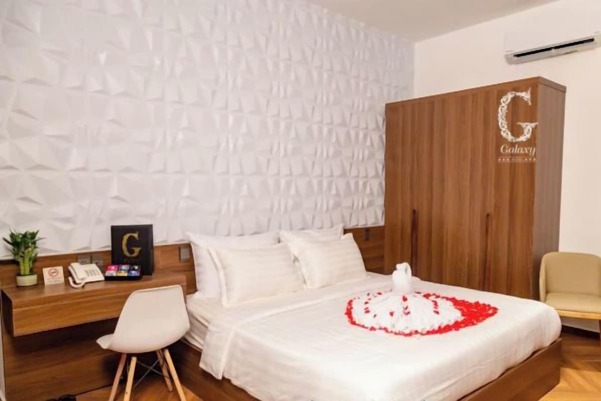 A hotel room with a bed, a wooden wardrobe, a desk with a phone, a chair, and a small table with a plant.