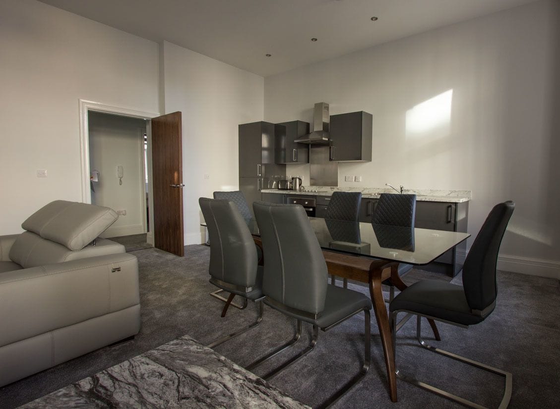 Una sala de estar moderna con un sofá gris, una mesa de comedor con seis sillas y una cocina con armarios oscuros y electrodomésticos de acero inoxidable.