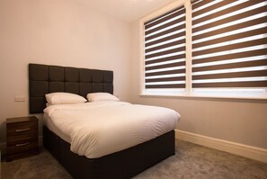 Een slaapkamer met een donker hoofdbord, witte beddengoed, een klein houten nachtkastje en een raam met horizontale luiken.