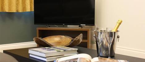 Un salon avec un téléviseur à écran plat, un saladier en bois, une bouteille de champagne et des magazines sur une table basse.