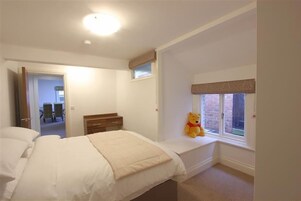 Un dormitorio con cama, un asiento junto a la ventana con un osito de peluche, y una puerta que da a otra habitación.