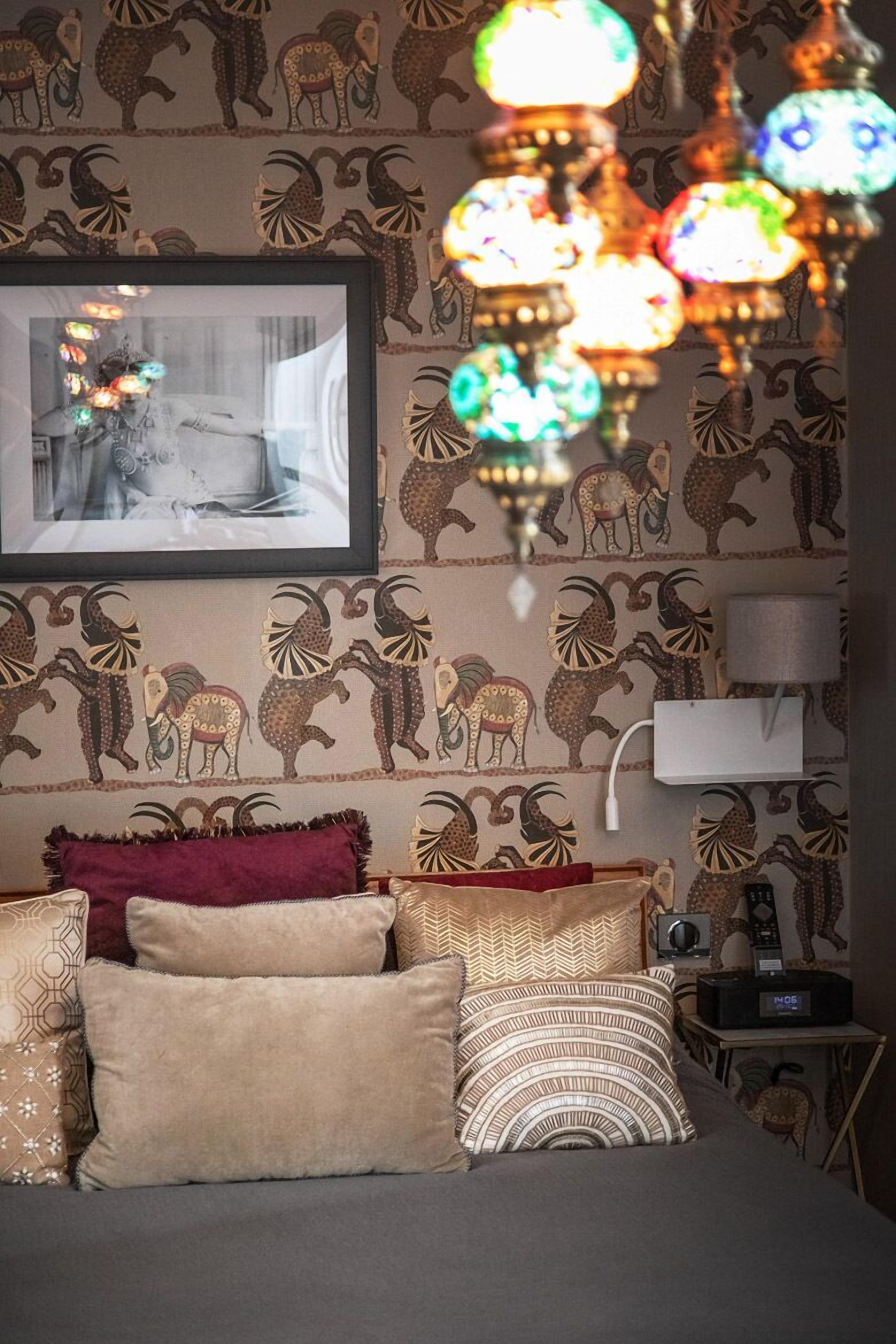 Une chambre avec un papier peint à motifs, un lit avec de nombreux coussins, un tableau encadré au mur, une lampe de chevet et une radio.
