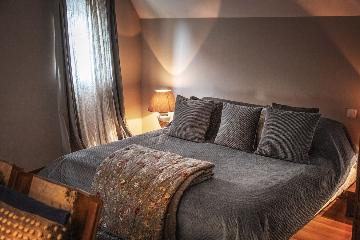 A neatly made bed with a grey quilt and pillows, a bedside lamp, and a window with curtains.