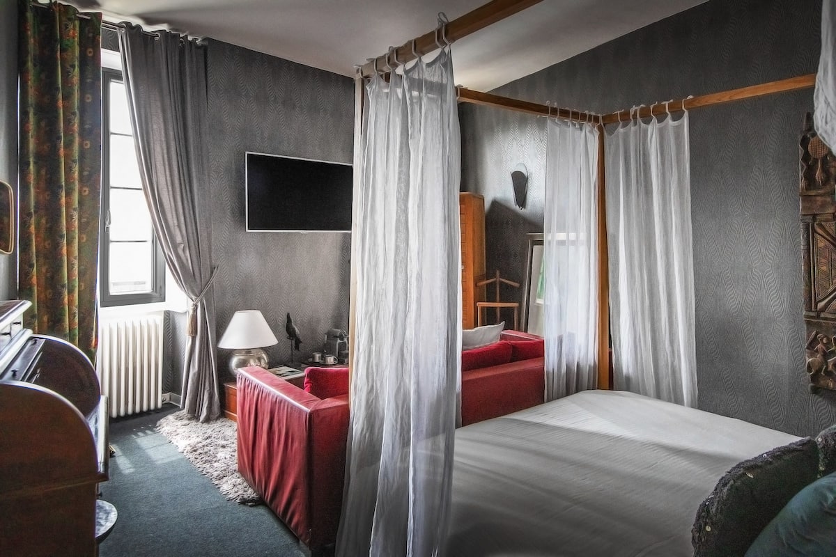 Une chambre à coucher avec un lit à baldaquin, un canapé rouge, un bureau avec une lampe et une grande fenêtre avec des rideaux.