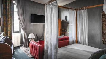 A bedroom with a four-poster bed, a red sofa, a desk with a lamp, and a large window with curtains.