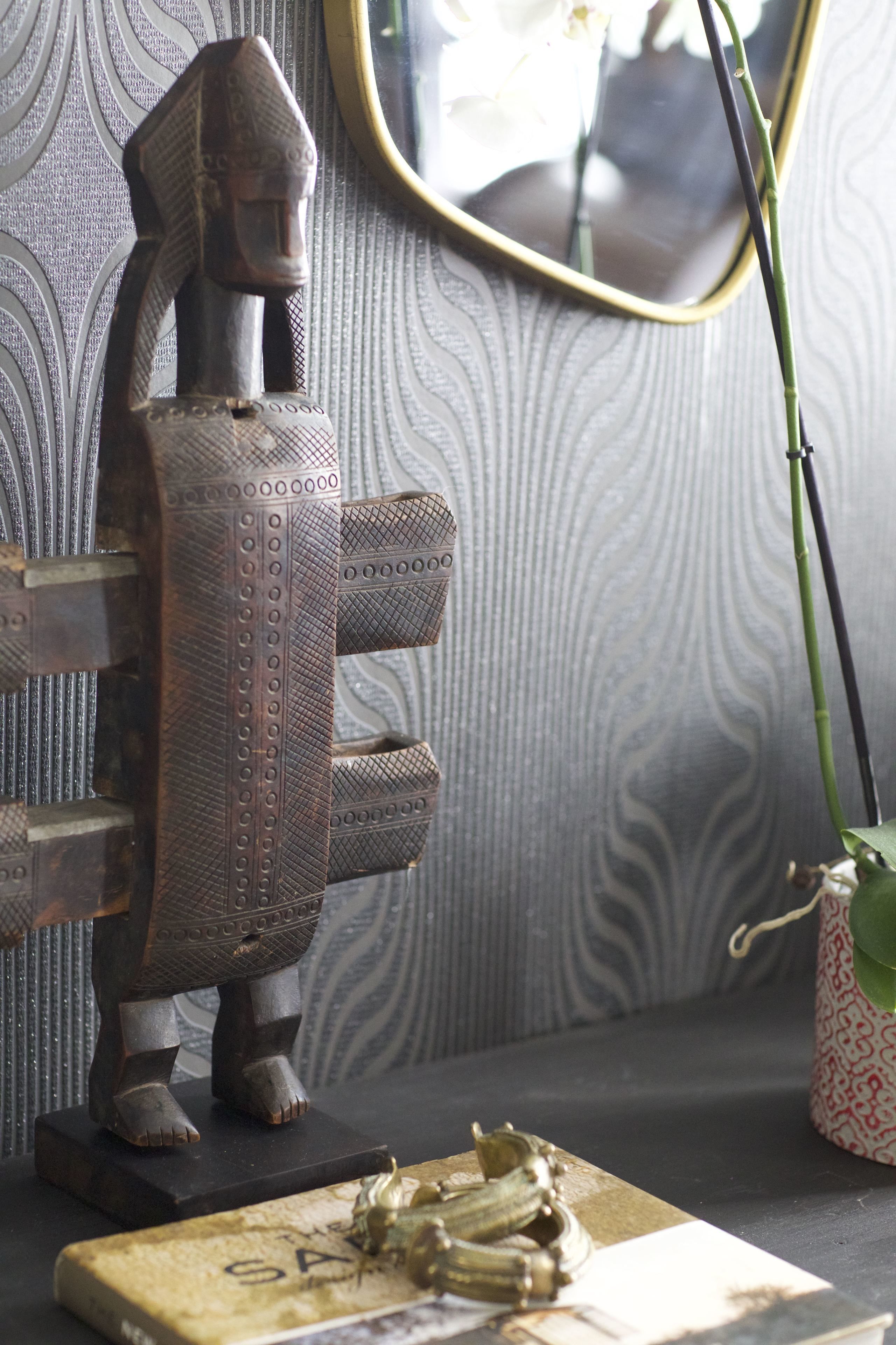 Een houten sculptuur op een zwarte voet, een patroonwand en een spiegel met gouden lijst.