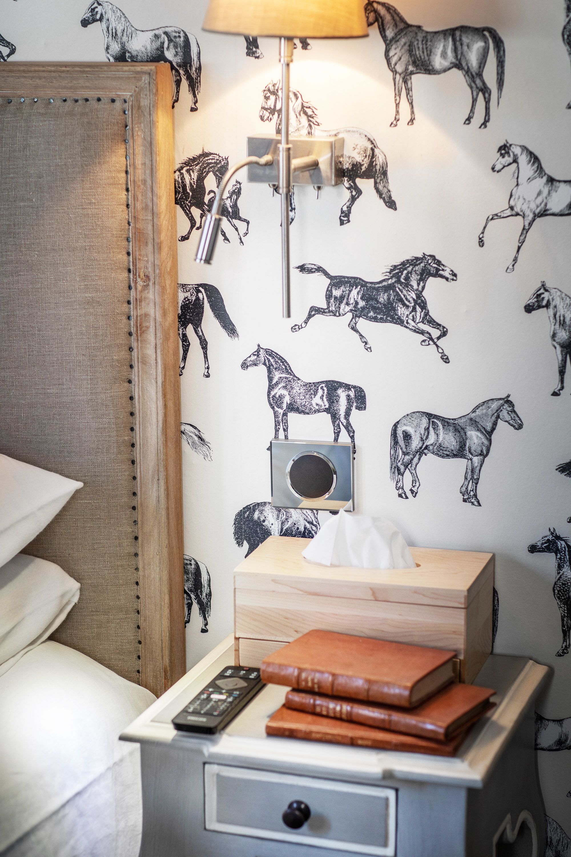 A bedroom with a bed, a bedside table with a lamp, a radio, and a stack of books.