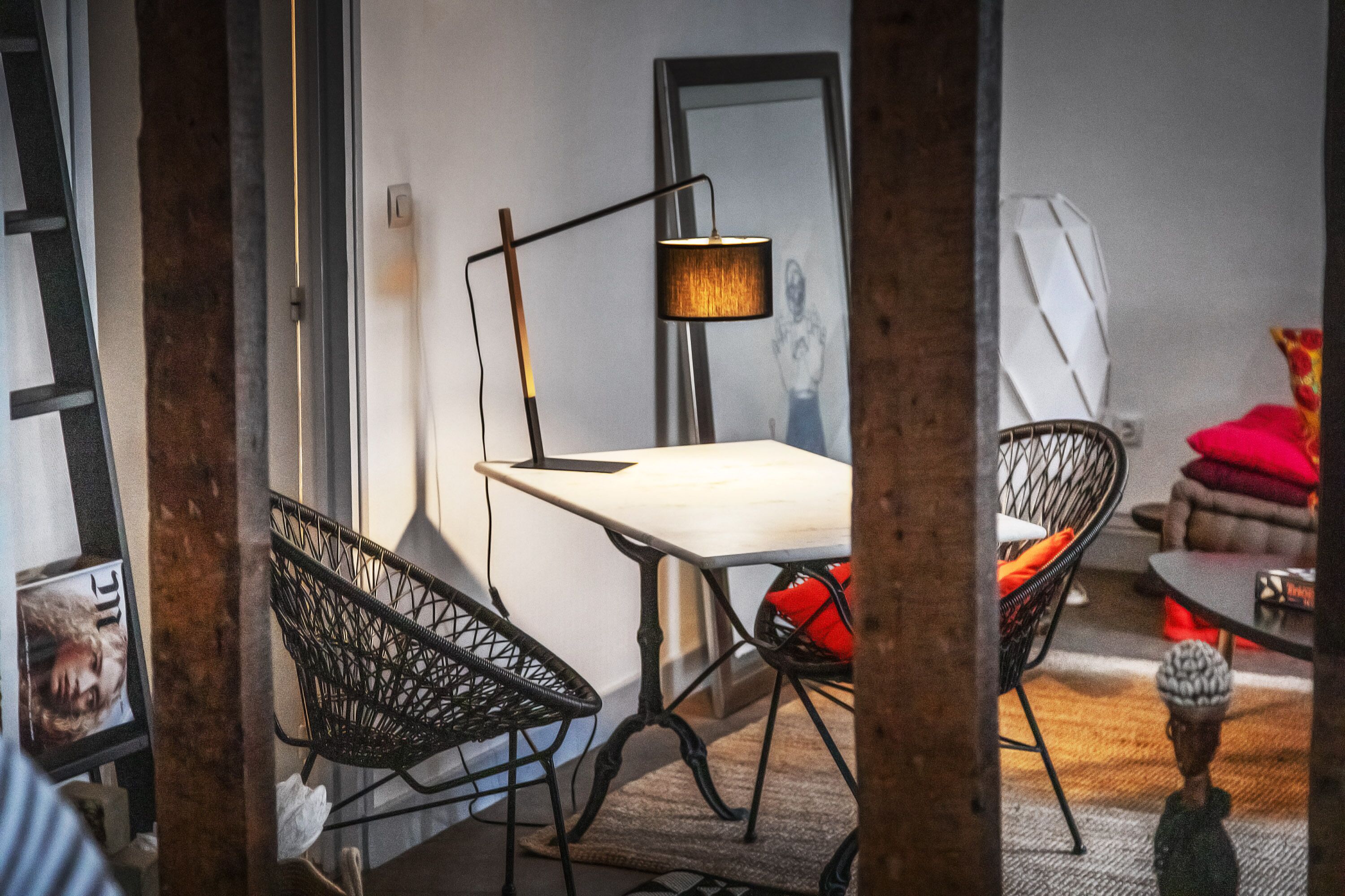 A modern living room with a white table, black wireframe chairs, a floor lamp, and a red cushion.