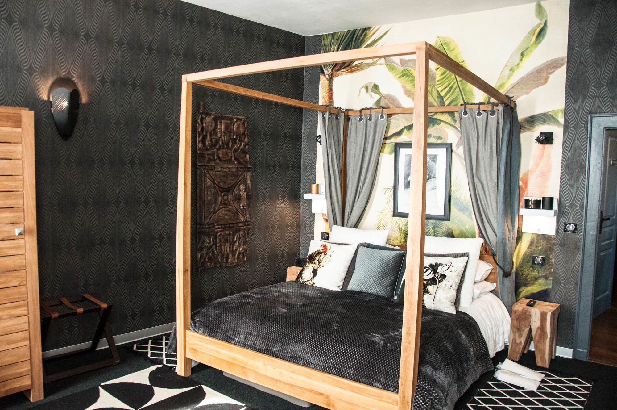 A four-poster bed with a dark, textured bedspread, a wooden headboard, and grey curtains.