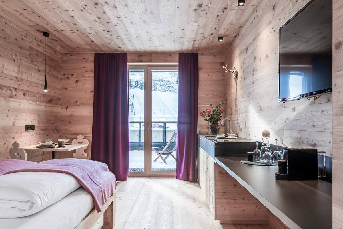 A wooden cabin room with a bed, a small table, a chair, a sink, and a television.