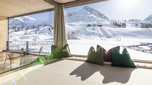 Een kamer met een groot raam met uitzicht op een besneeuwd berglandschap, een houten plafond en een zithoek met kussens.
