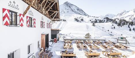 Un área de estar al aire libre cubierta de nieve con mesas y bancos de madera, un edificio con señalización roja y blanca, y un paisaje montañoso de fondo.