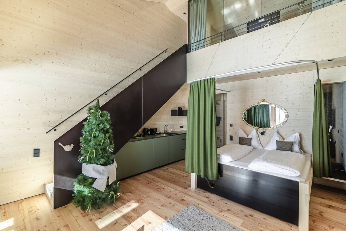A modern loft bedroom with a large bed, a small green Christmas tree, a kitchen area, and a staircase.